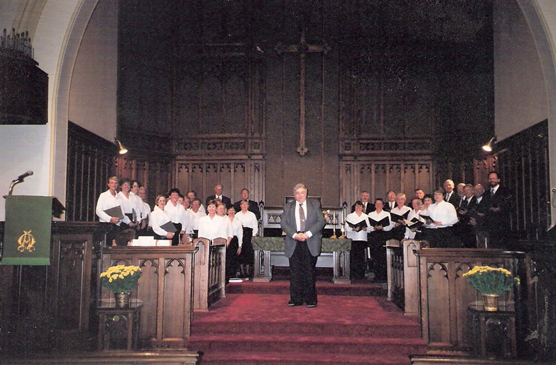 A choir standing in the choir section of a church, with a man at the centre. Choir members are wearing black and white. 